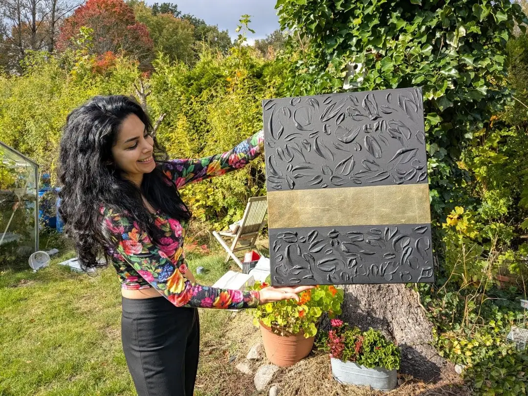 Mona i profil i en trädgård, betraktar ett grått texturerat verk med ett guldband som hon håller upp framför häcken.